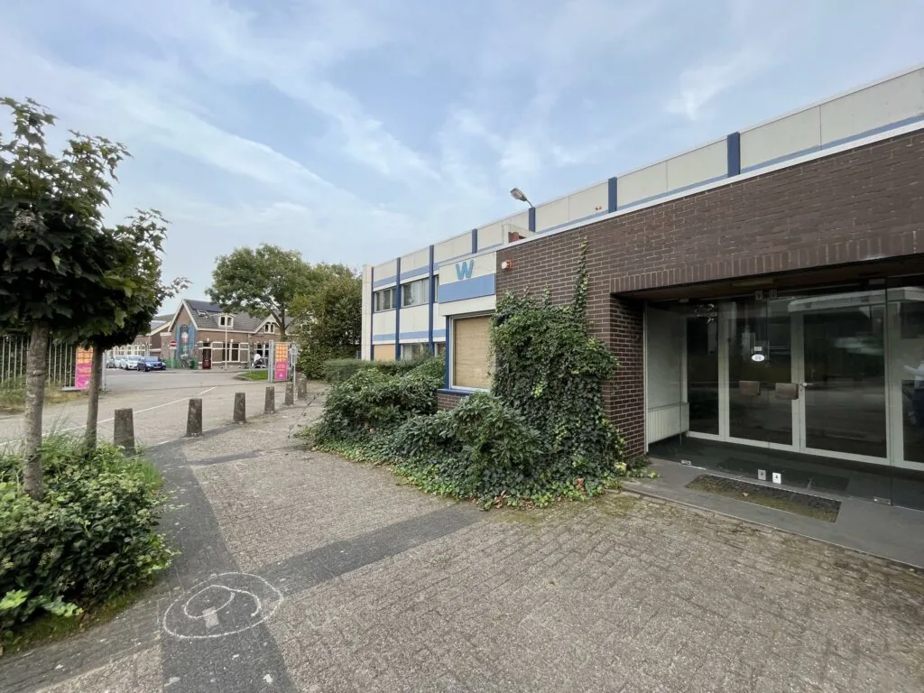 Entree van een leegstaand bedrijfsgebouw aan de 2e Daalsedijk met een betegeld pad en overwoekerde planten.